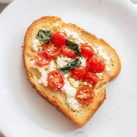 Overhead view of four slices of baked feta toast on a parchment-lined tray, with blistered tomatoes and glistening olive oil for a Mediterranean-inspired snack.