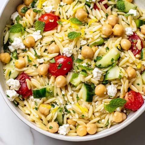 A close-up of Lemony Chickpea Orzo Salad in a white bowl, showcasing tender orzo, chickpeas, diced cucumber, and cherry tomatoes tossed in a bright lemon vinaigrette.  